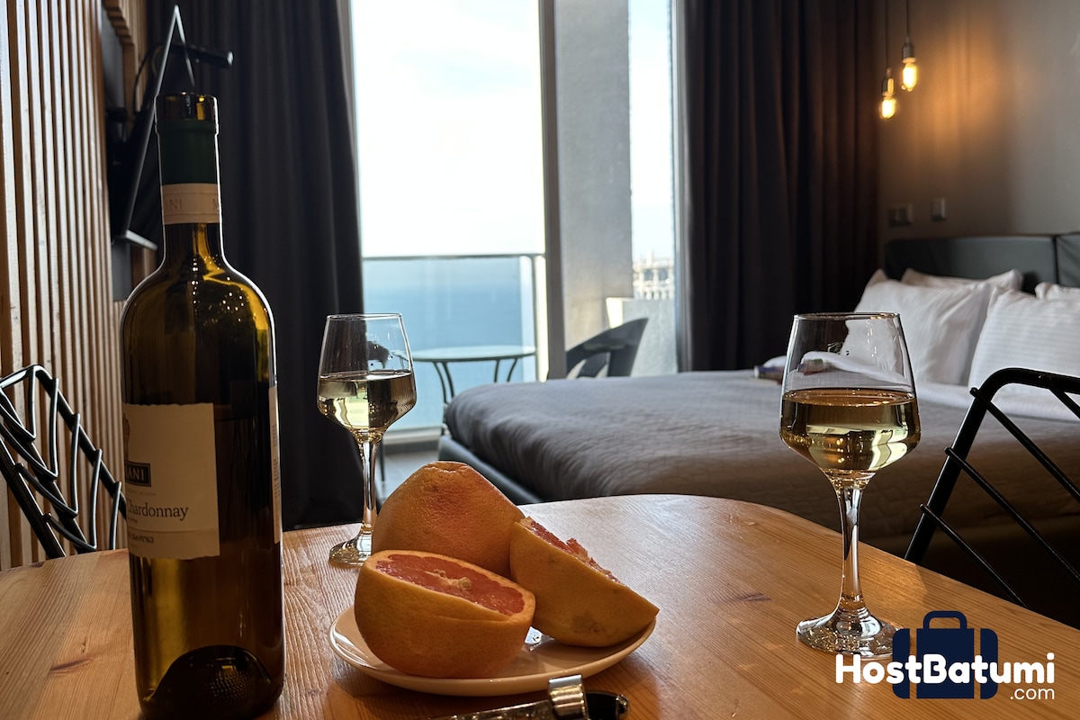 A table is set with two glasses of white wine and fresh fruit, creating an inviting dining scene. In the background, a spacious bed is visible, along with large windows that reveal a sea view, allowing natural light to brighten the space.
