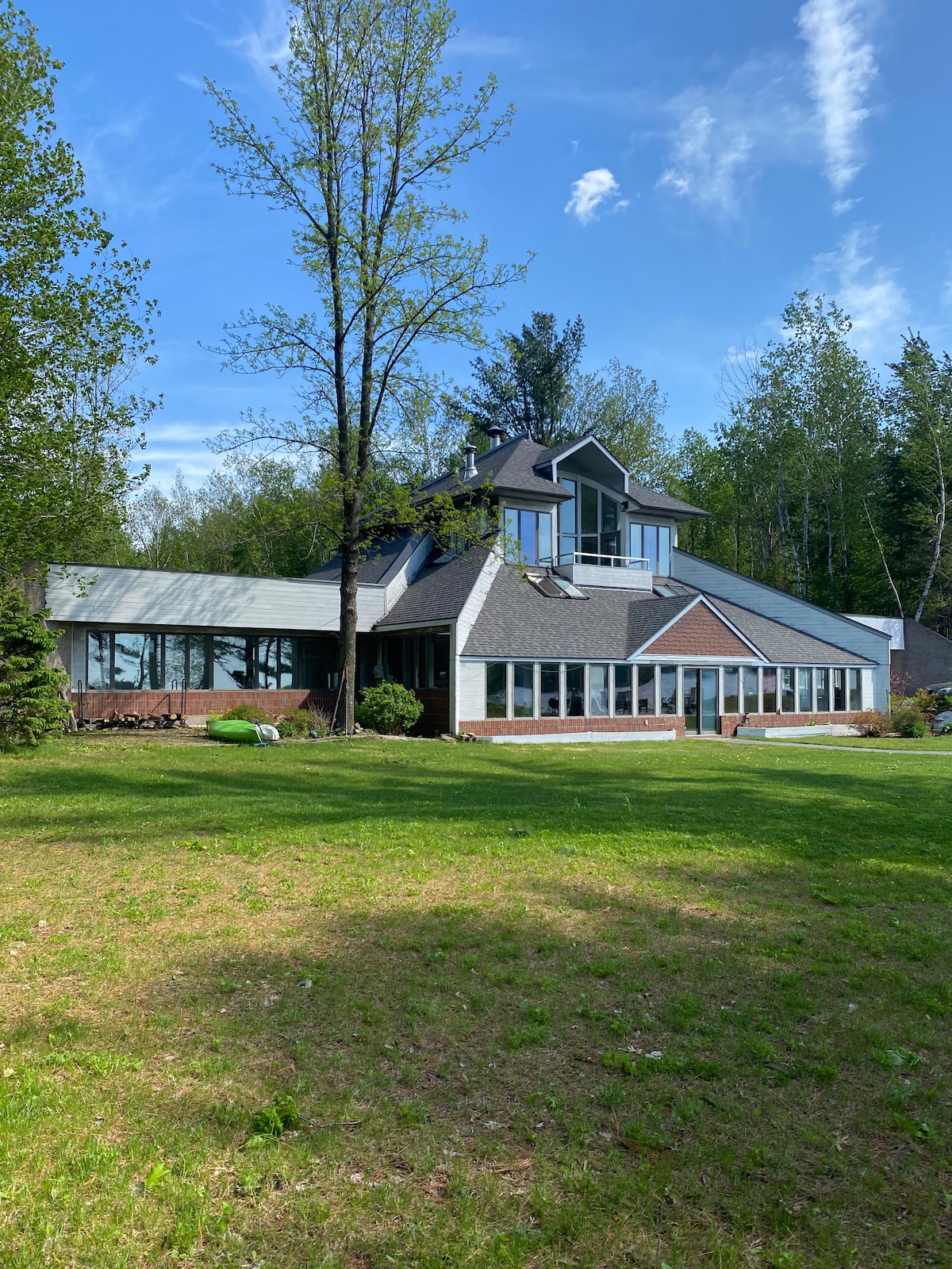 The exterior of a spacious hilltop home is shown, surrounded by a grassy area and mature trees. A wall of windows highlights the house's design, allowing for natural light. The structure features a mix of brick and siding, complemented by a central peaked roof.