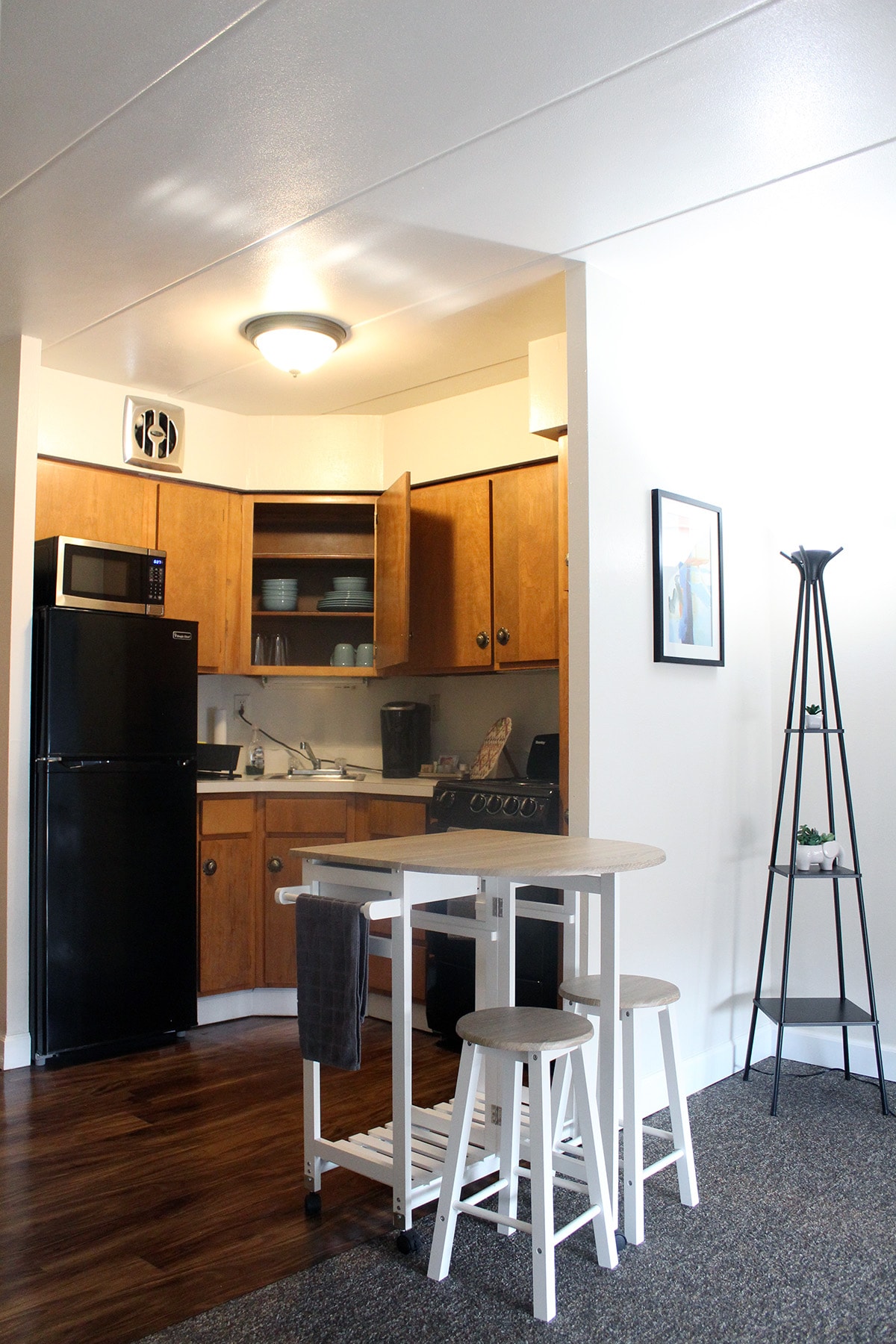 A compact kitchen area is highlighted, featuring wooden cabinetry and essential appliances including a refrigerator and microwave. A small dining table with three stools occupies the space, while a decorative stand supports a plant in the corner, adding a touch of greenery.