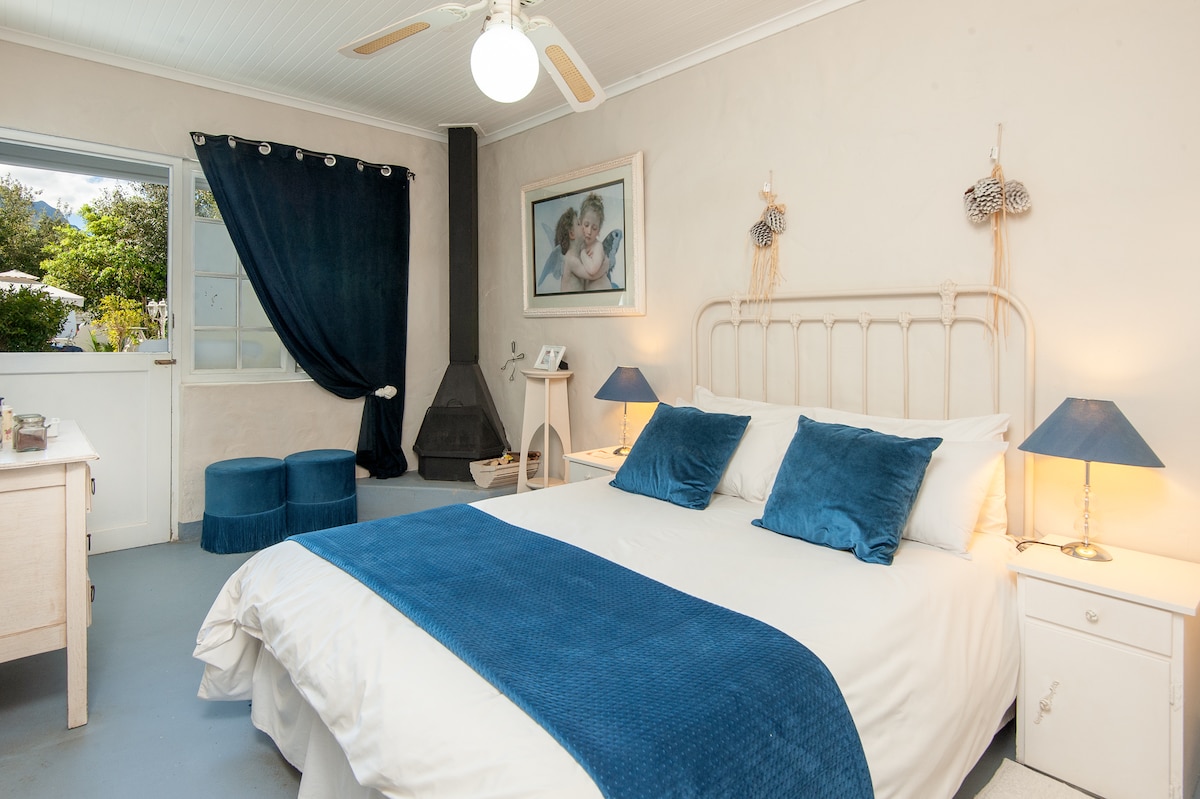 A well-decorated bedroom features a spacious bed dressed in white linens with blue accents. Two bedside lamps provide soft lighting. A wood stove stands nearby, and a window showcases natural light filtered through dark curtains, offering insights into the room's tranquil atmosphere.