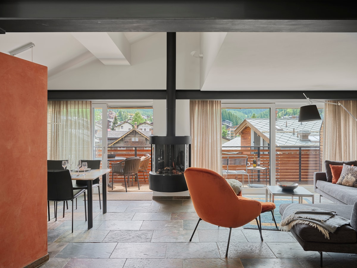 A bright living area features a modern fireplace at the center, surrounded by large glass doors that open to a balcony. An orange chair adds a pop of color against the neutral tones, while a dining table is set for four, creating a welcoming space.