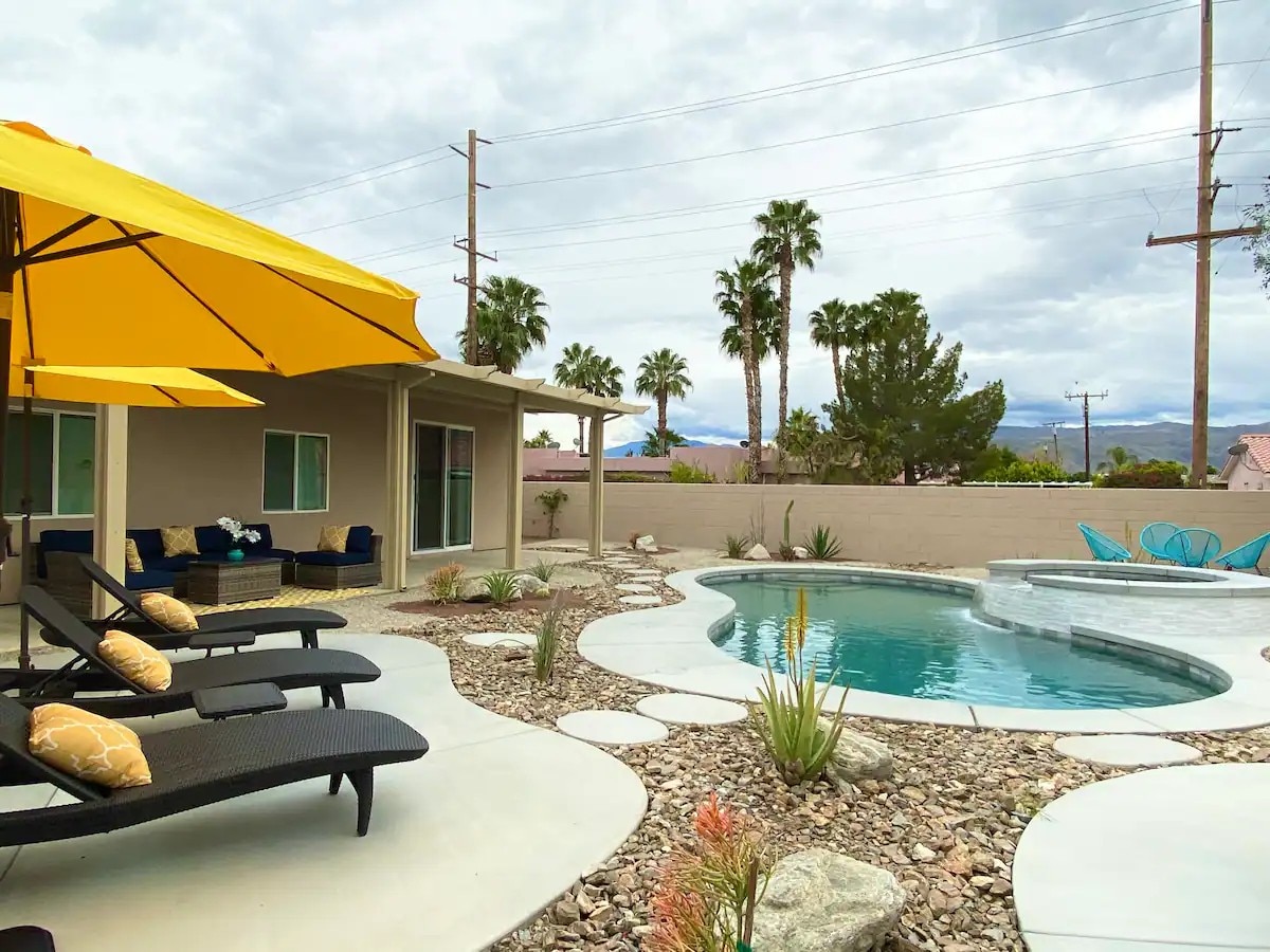 An outdoor seating area features yellow umbrellas providing shade over lounge chairs. A swimming pool is surrounded by landscaped stones and desert plants. Palm trees are visible in the background, with cloudy skies creating a tranquil atmosphere.