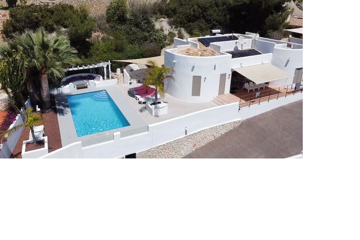 An aerial view of a modern villa showcases a private pool surrounded by a spacious outdoor deck. Lush greenery borders the property, providing privacy. The villa features large windows and outdoor seating areas for relaxation. The sunlit setting enhances the tranquil atmosphere of the surroundings.