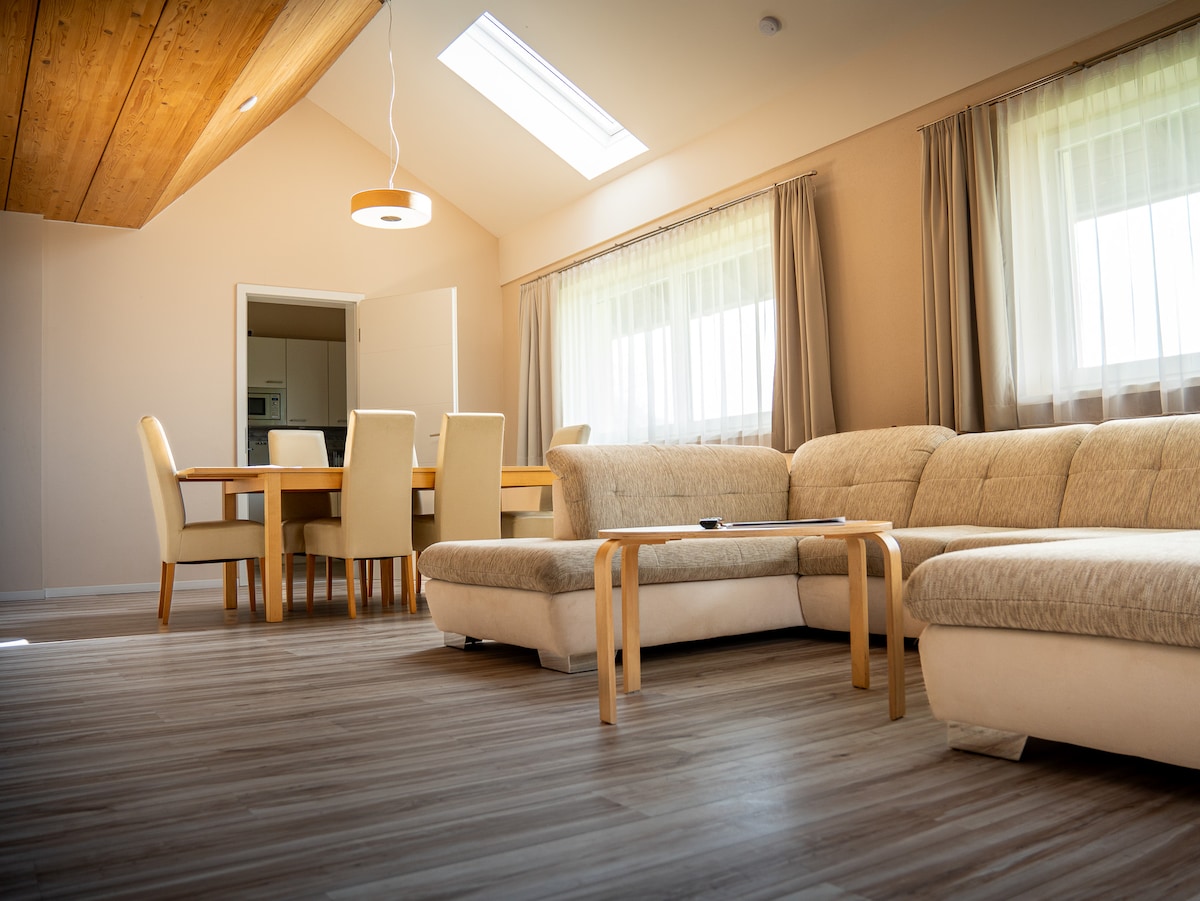 A spacious living area features a light-colored sectional sofa and a wooden coffee table, providing comfort for relaxation. Natural light filters through large windows covered with sheer curtains. A dining table with light-colored chairs is visible in the background, enhancing the overall openness of the room.