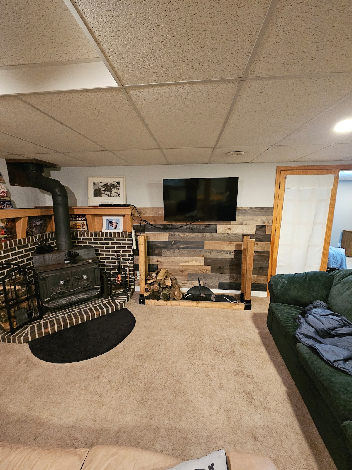 A living area features a cozy wood fireplace, surrounded by stacked logs. A mounted television is positioned on a wooden wall, complemented by various framed photos. A plush green sofa is available for seating, with soft carpet underfoot and a subtle light illuminating the space.