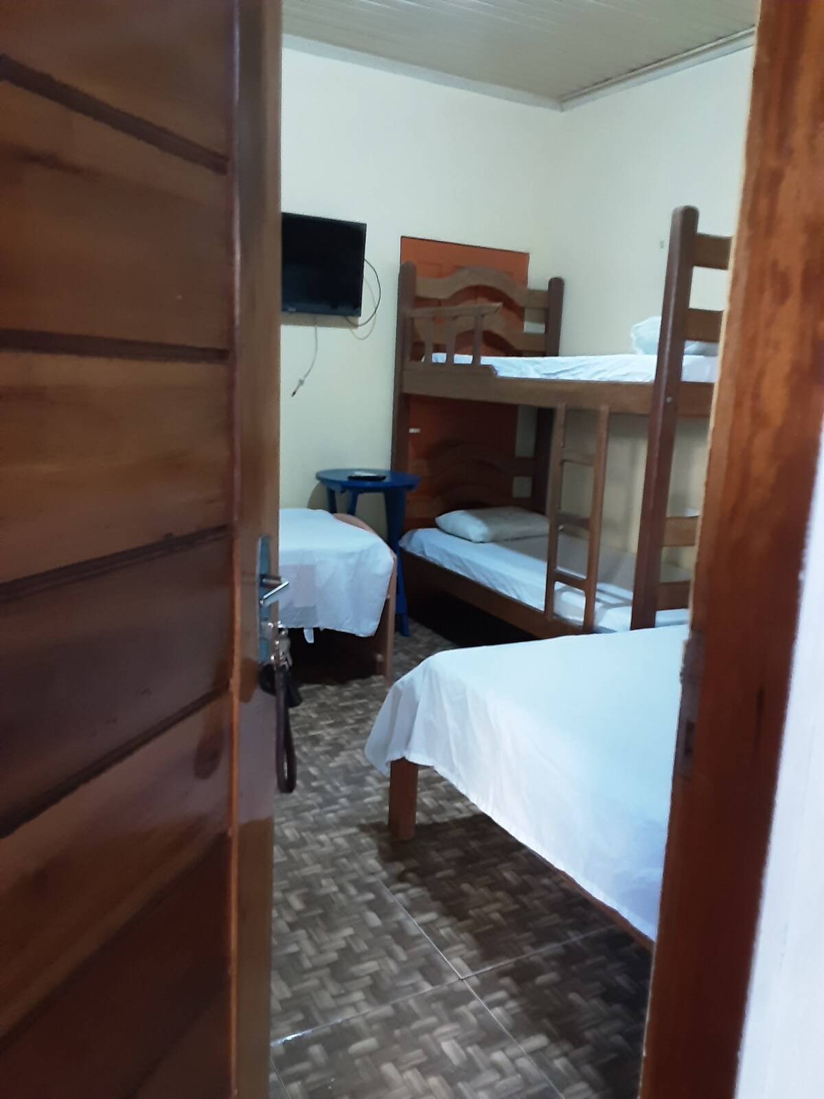 A bedroom featuring a bunk bed and a single bed, surrounded by wooden walls. A small nightstand is placed beside the single bed. A television is mounted on the wall, while natural light enters through the doorway.