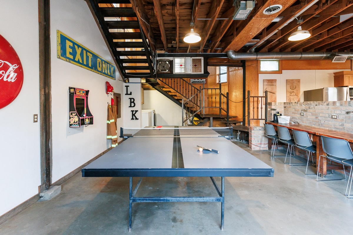 A spacious game room features a large black ping pong table at its center, with retro arcade games visible against the wall. The warm wooden beams and open staircase create an inviting and unique atmosphere. A well-equipped kitchen is visible in the background.