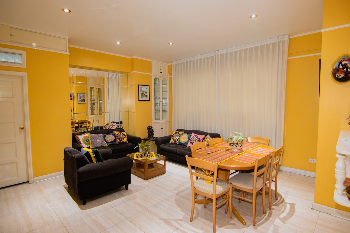 A spacious living area features vibrant yellow walls and stylish furnishings. Two dark sofas adorned with colorful cushions are positioned around a coffee table. A light-colored dining table with wooden chairs is set beside a large window, offering ample natural light.