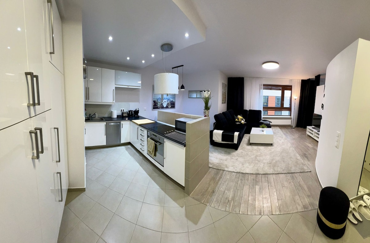 A modern kitchen and living area are visible, featuring sleek white cabinetry and stainless steel appliances. The spacious living room offers a comfortable black sofa facing a flat-screen TV. Natural light enters through large windows, enhancing the clean and contemporary design of the space.