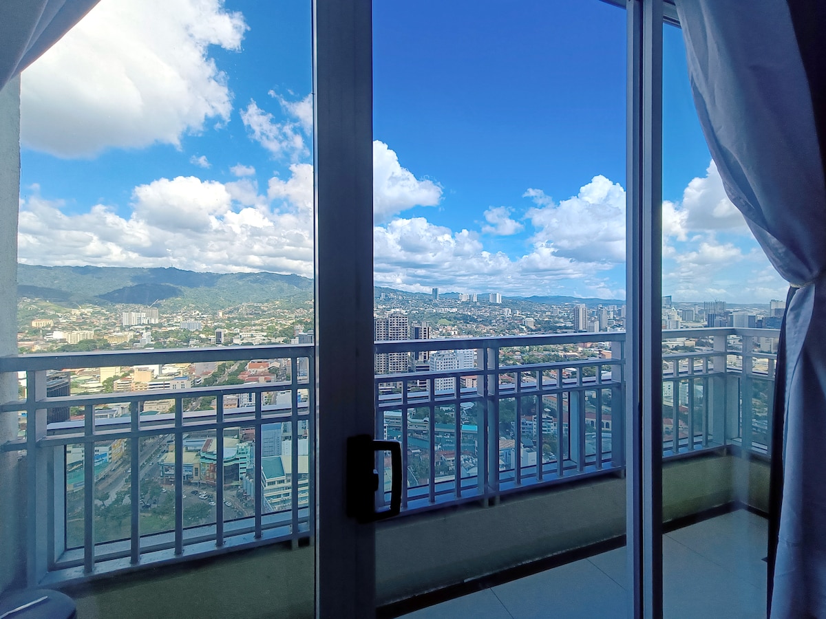 An expansive view of Cebu is presented from the penthouse balcony, showcasing a vibrant skyline amid lush mountains. The scene captures a bright blue sky dotted with fluffy white clouds, offering a serene backdrop for relaxation.