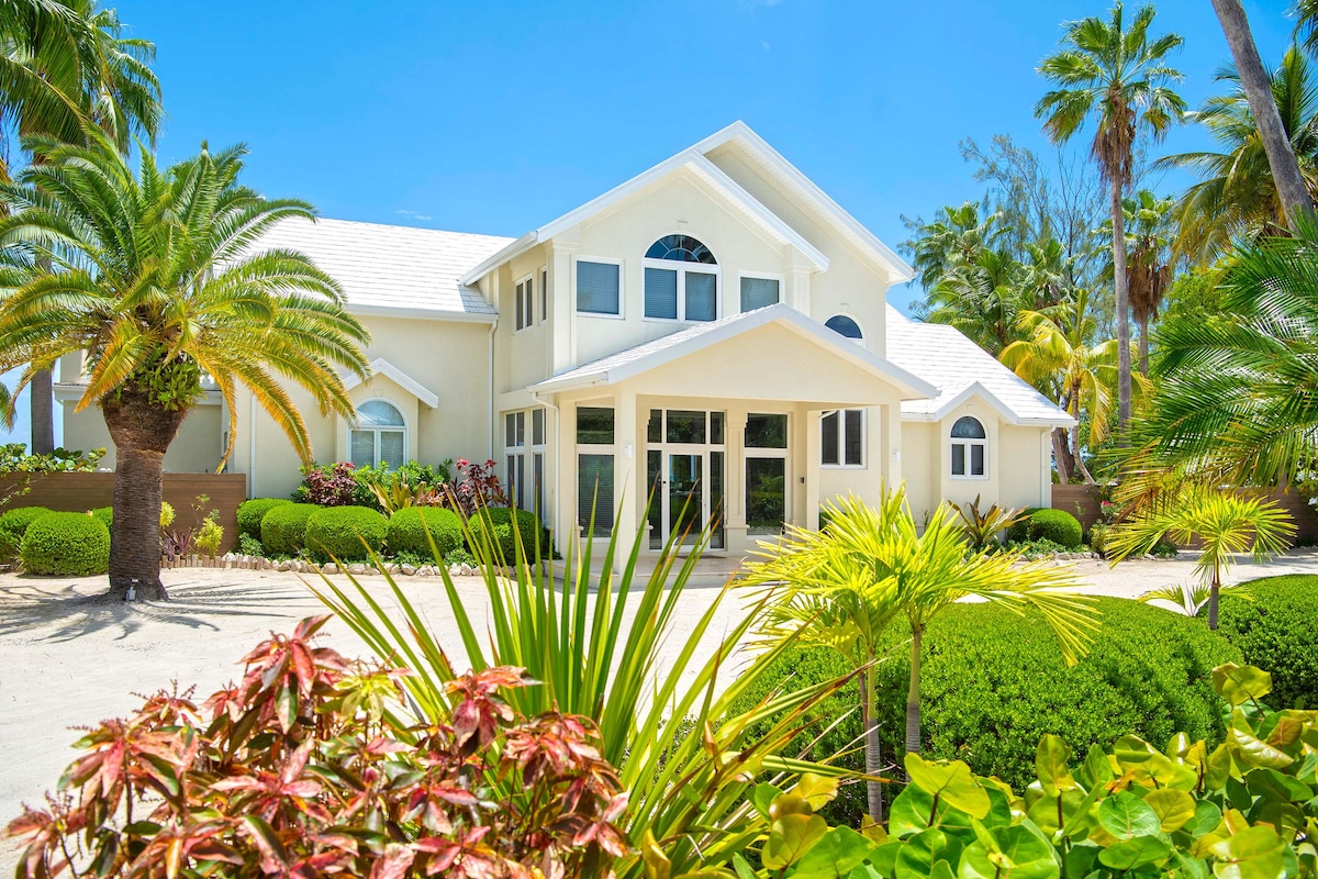 The villa's exterior features a welcoming entrance framed by lush tropical landscaping. Large windows allow natural light to flood the interior, while palm trees sway gently in the breeze, creating a serene atmosphere against the bright blue sky.