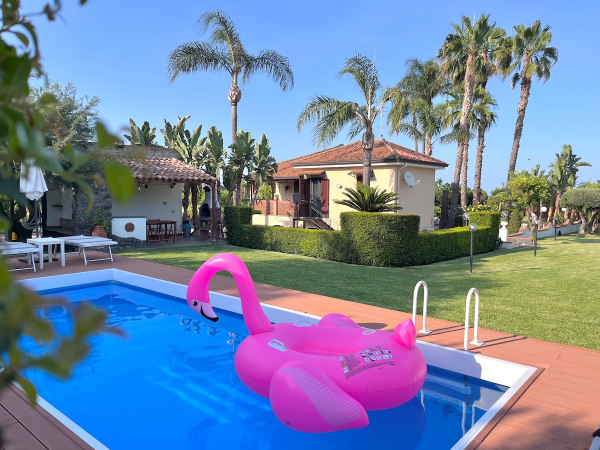 A serene outdoor area features a private pool with a pink inflatable float. Surrounded by lush greenery, palm trees, and a well-maintained garden, the space invites relaxation under the clear blue sky. The villa, with its traditional roof, sits gracefully in the background.