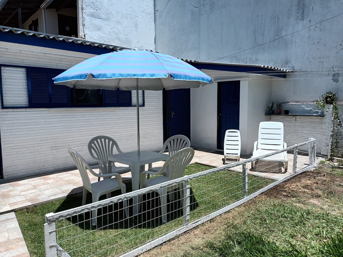 A shaded outdoor area features a round table under a blue-striped umbrella, accompanied by four white chairs. A separate lounge chair is positioned nearby on well-maintained grass. The backdrop includes a small building with blue doors and windows, contributing to a welcoming ambiance.