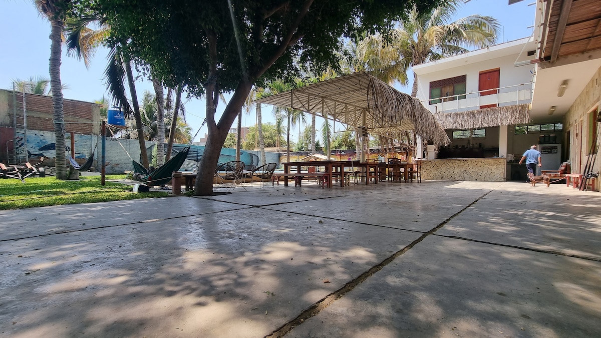 An expansive outdoor area features a concrete surface shaded by trees. A thatched roof covers a large gathering space with dining tables. Comfortable hammocks and palm trees enhance the relaxing environment. Nearby, a well-maintained lawn adds greenery to the scene.