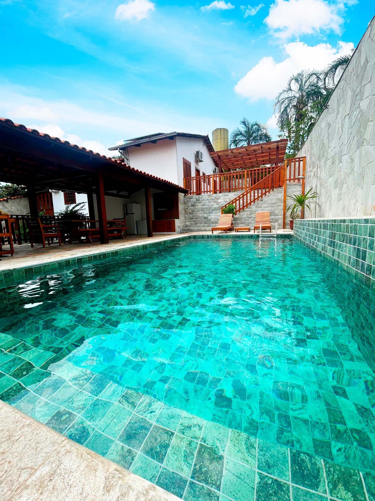 A clear, inviting pool reflects the blue sky and surrounding greenery, framed by natural stone and tiled walls. Adjacent areas include a shaded outdoor dining space and wooden deck, providing a scenic spot for relaxation.