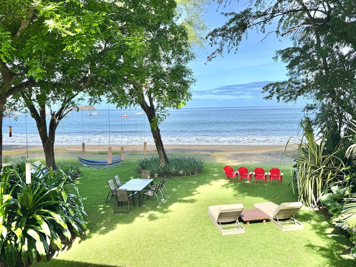 A serene beachfront garden is depicted, featuring a grassy area with lounge chairs and a dining table. The ocean is visible in the background, framed by lush trees. A hammock hangs nearby, while vibrant red outdoor chairs add a pop of color to the setting.