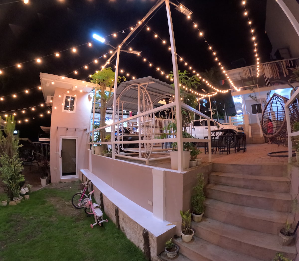The outdoor area is illuminated with string lights, creating a cozy ambiance. Comfortable seating areas are visible, along with a swing and decorative plants. A small grassy section is seen at the base of the stairs that lead to the patio.