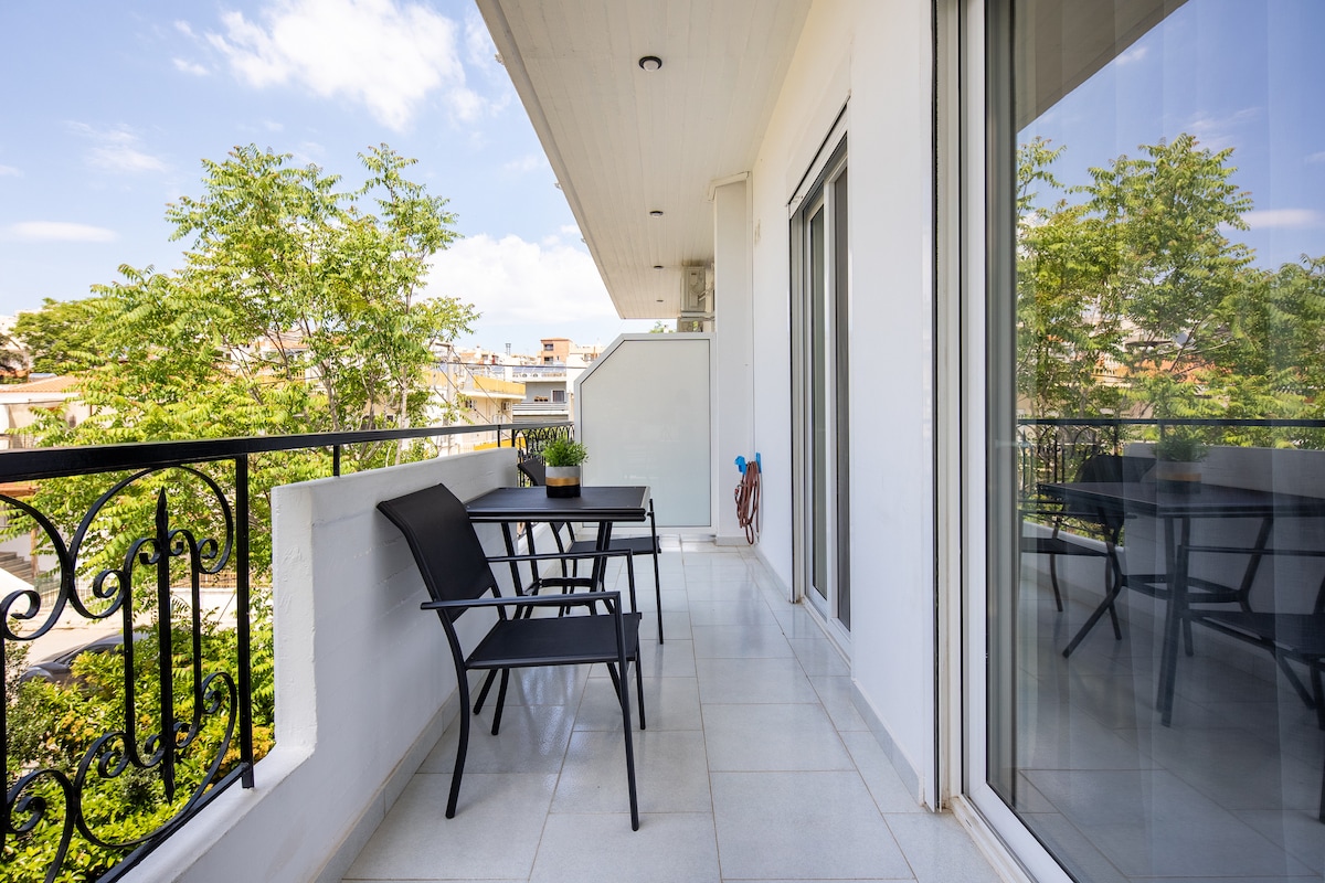 A spacious balcony features a table with two chairs, surrounded by greenery visible from the lower level. Large sliding glass doors connect the interior space to the balcony, allowing for natural light to flow into the area.