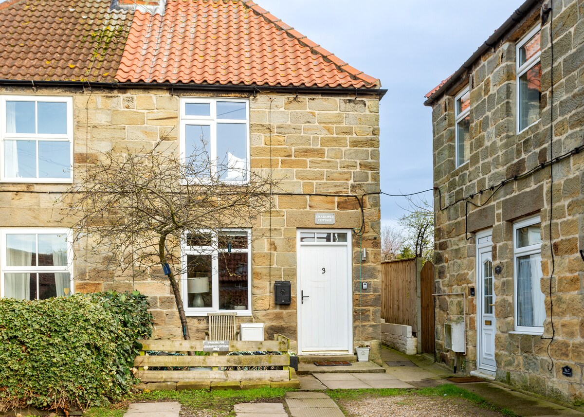 The exterior of Crabapple Cottage is shown, featuring a traditional stone façade and a welcoming white front door. A small bench is situated on a stone path, surrounded by greenery. The cottage is nestled in a cozy courtyard with neighboring stone buildings visible nearby.