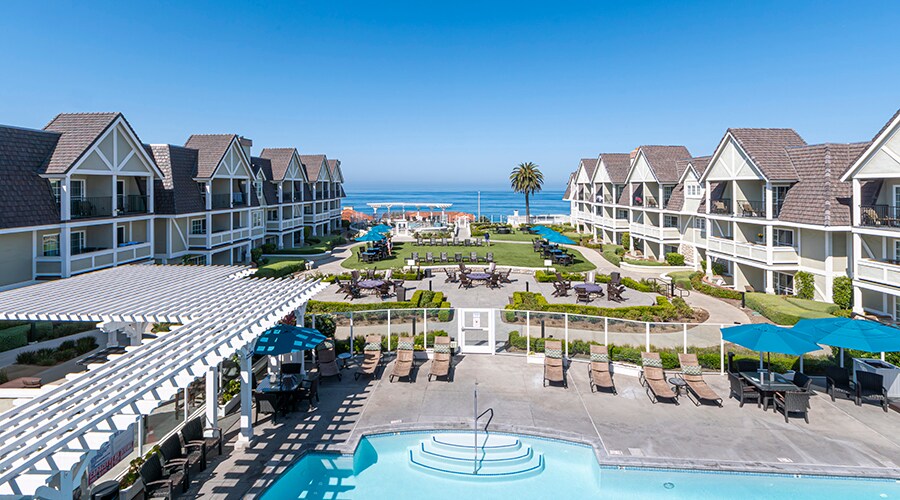 The view showcases the resort's beautifully landscaped grounds, featuring a large outdoor pool and seating areas. Beachfront cabanas and loungers are arranged neatly under shade. The Pacific Ocean glimmers in the background, framed by the resort's charming architecture.