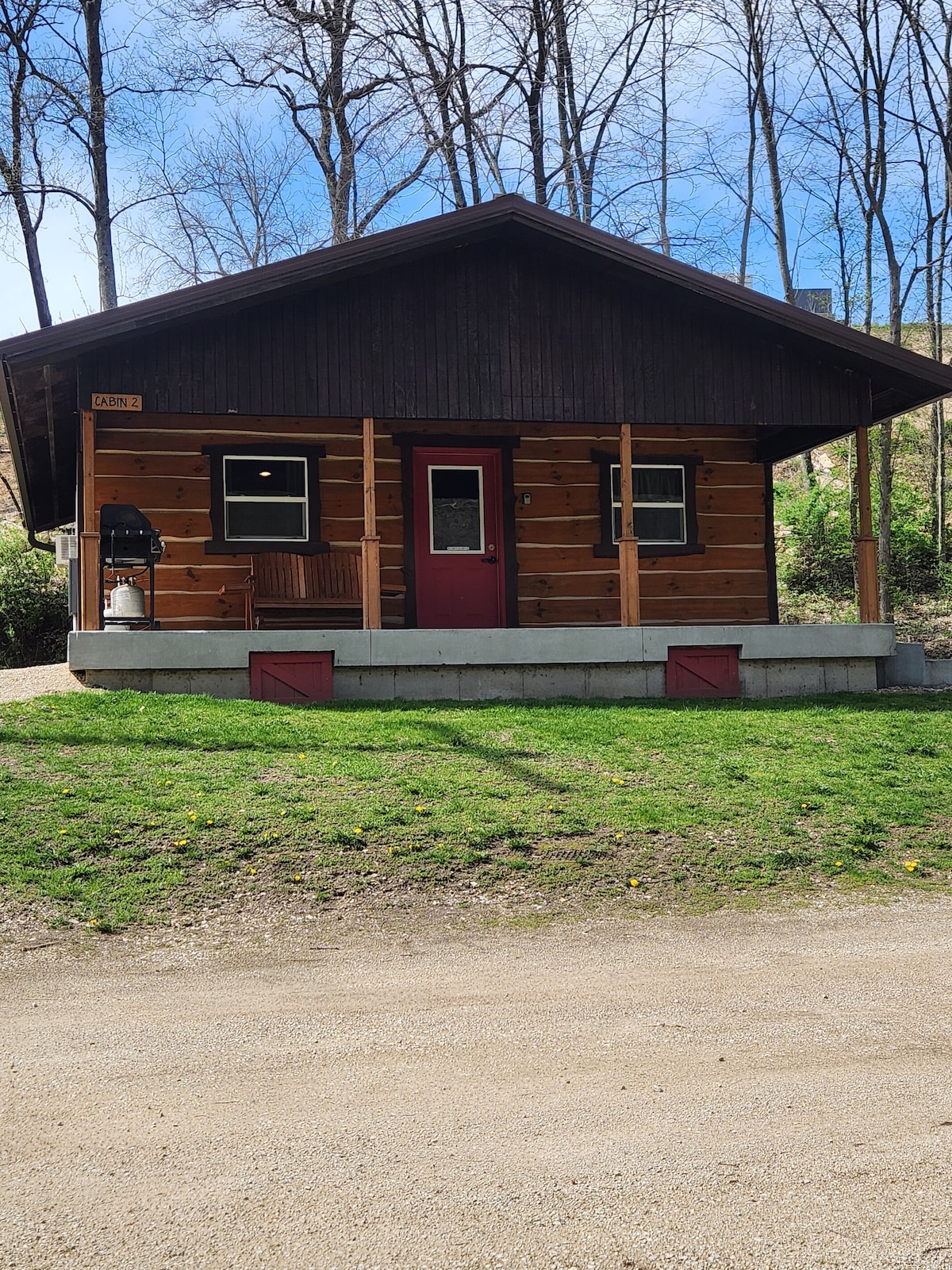 The cabin features a rustic wooden exterior with dark accents and a welcoming red door. A covered porch provides seating, and small windows allow natural light to brighten the space. The surrounding green grass enhances the natural setting of the cabin.