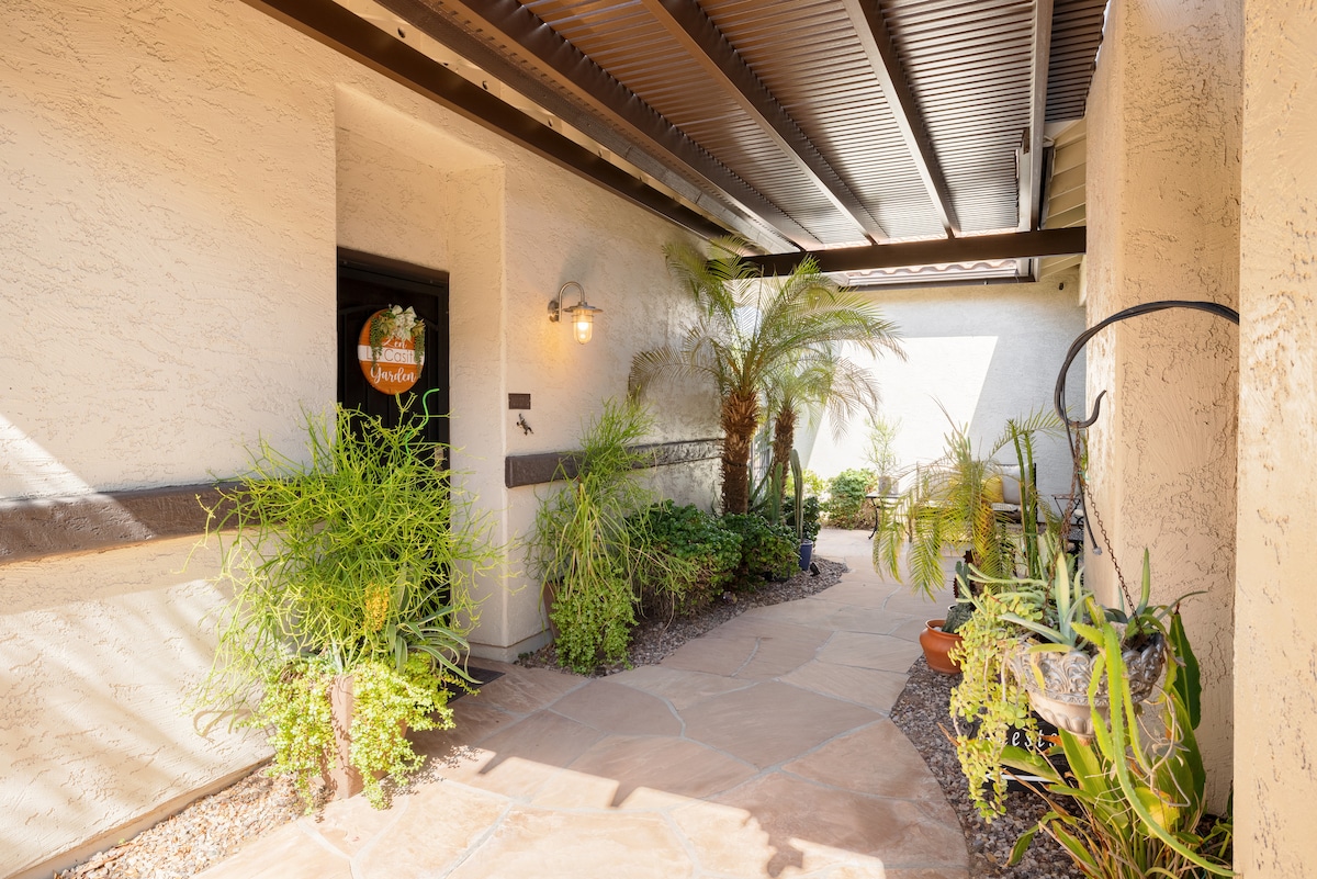 The entrance pathway is framed by lush greenery and soft landscaping. A stone walkway leads towards the entrance, with sunlight filtering through the covered patio. Plants in pots and a palm tree add a touch of nature to the welcoming atmosphere.