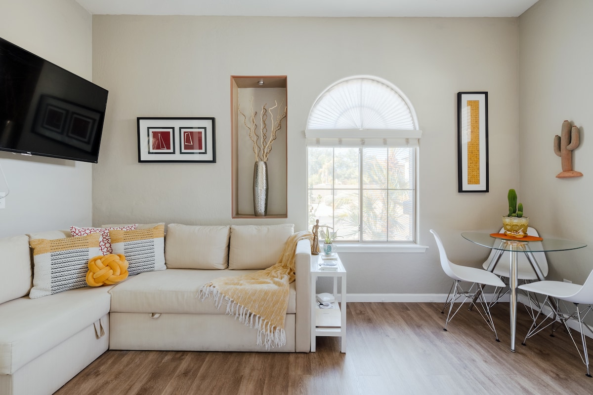 A bright and airy living space features a comfortable light-colored couch adorned with decorative pillows and a soft throw. A glass dining table for two is set beside a window with elegant window treatments, while wall art and decor add a touch of personality.