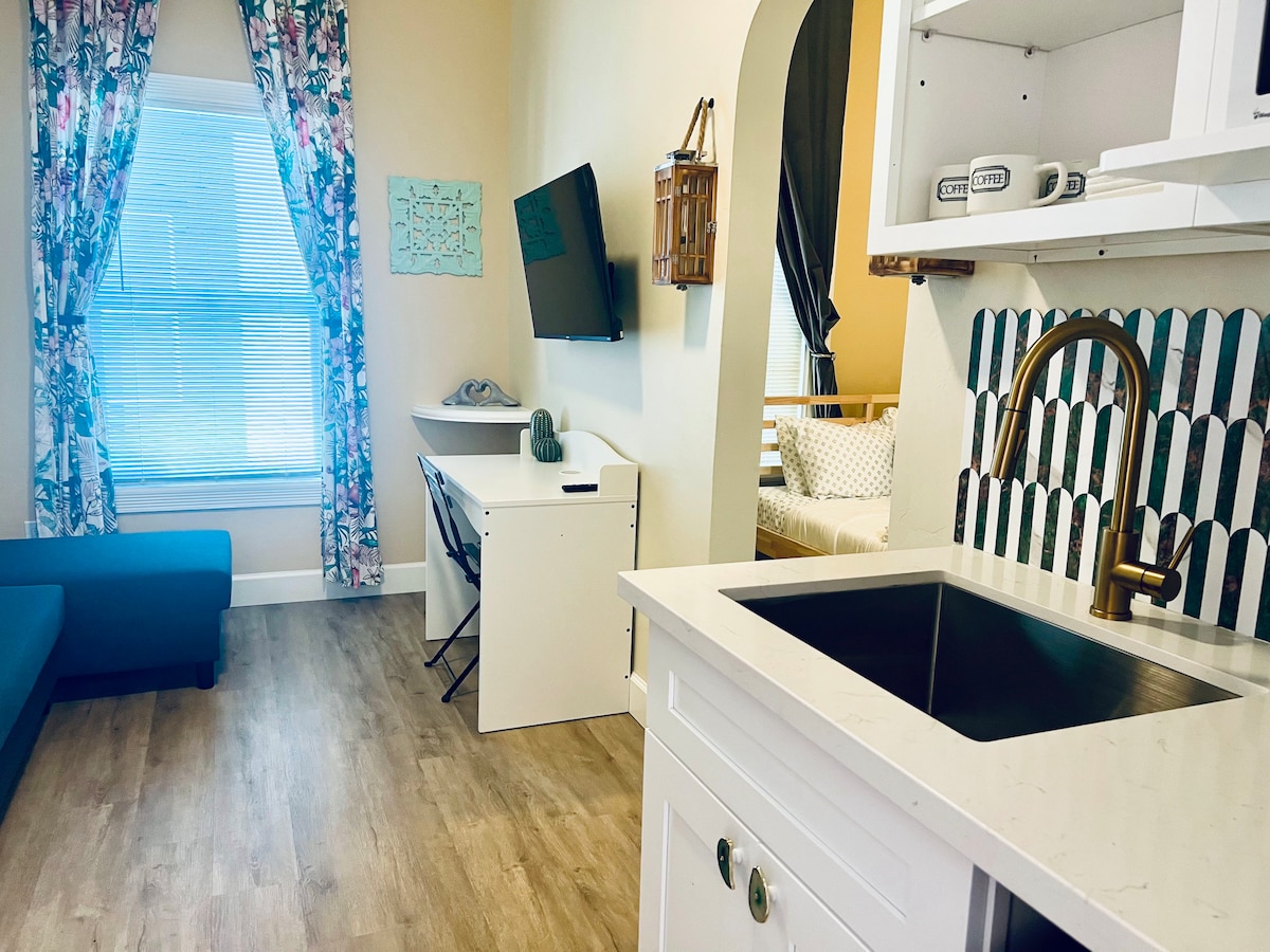 The room features a cozy seating area with a teal couch and a workspace with a white desk. A kitchenette area includes a sleek sink with a modern faucet, complemented by patterned backsplash. Large windows allow natural light to brighten the space, framed by floral curtains.