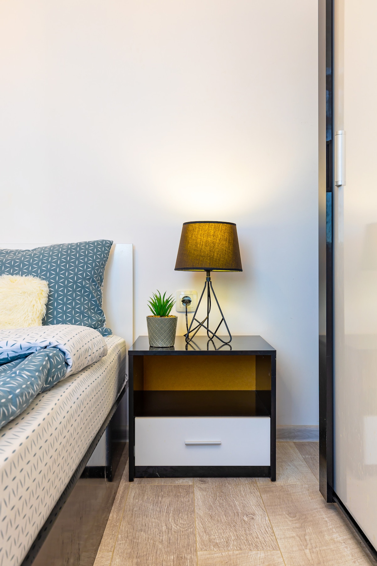 A bedside area is featured with a compact nightstand, which includes a small potted plant and a lamp. Soft lighting from the lamp creates a serene ambiance. The bed is dressed in a patterned duvet, with a textured throw resting atop. A wardrobe stands nearby.