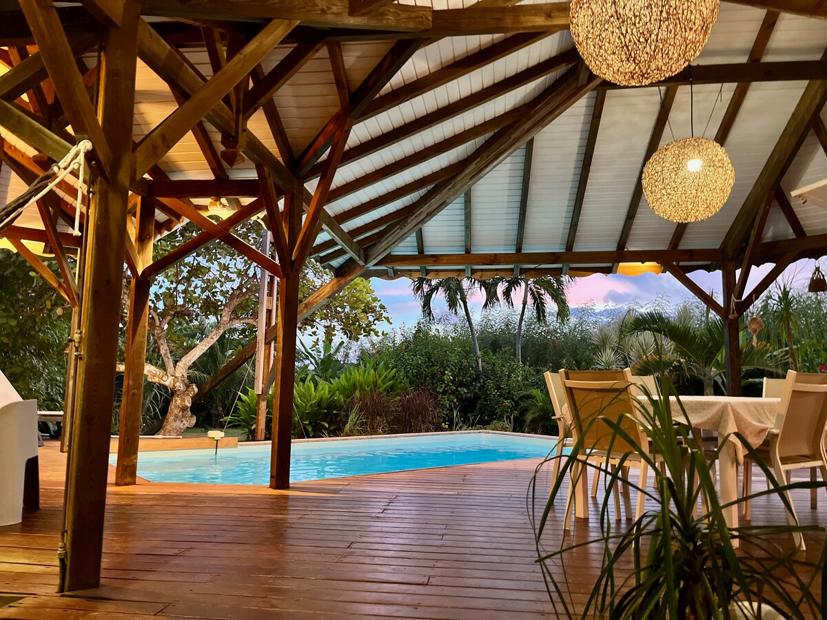 An inviting outdoor space is highlighted by a private swimming pool surrounded by a wooden deck. A rustic roof structure complements the area, with dining furniture and lush tropical greenery in the background. Soft lighting fixtures create a warm ambiance as dusk approaches.