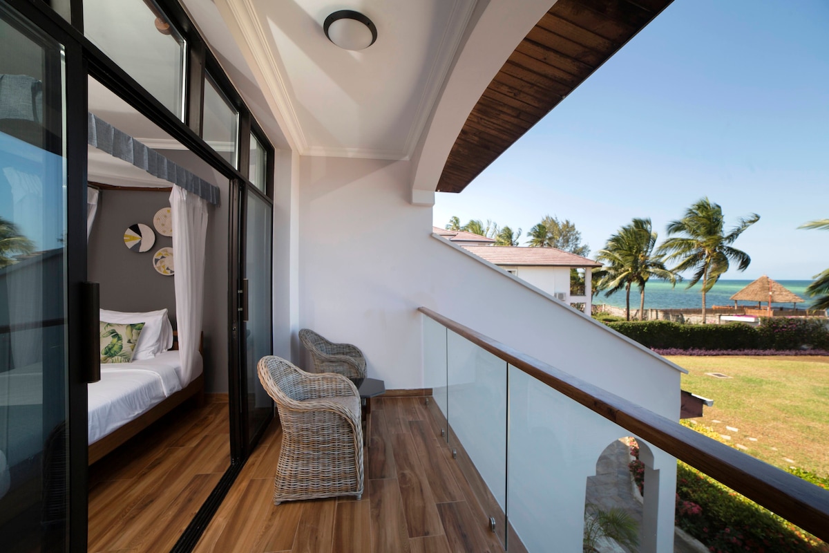 A private veranda features two wicker chairs, providing a comfortable space to enjoy ocean views. The glass doors lead to an interior room, while palm trees and the sea are visible in the distance, showcasing the tranquil surroundings.