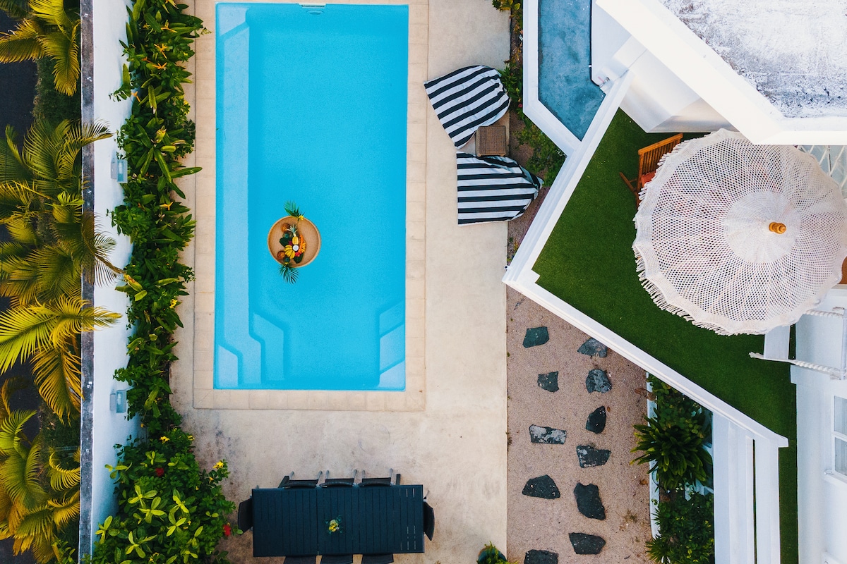 An aerial view showcases a private swimming pool surrounded by tropical plants and seating areas. Two striped lounge chairs are positioned near the pool, and a round table with decor sits at its center. The outdoor dining area features a long table, complemented by a sun umbrella.
