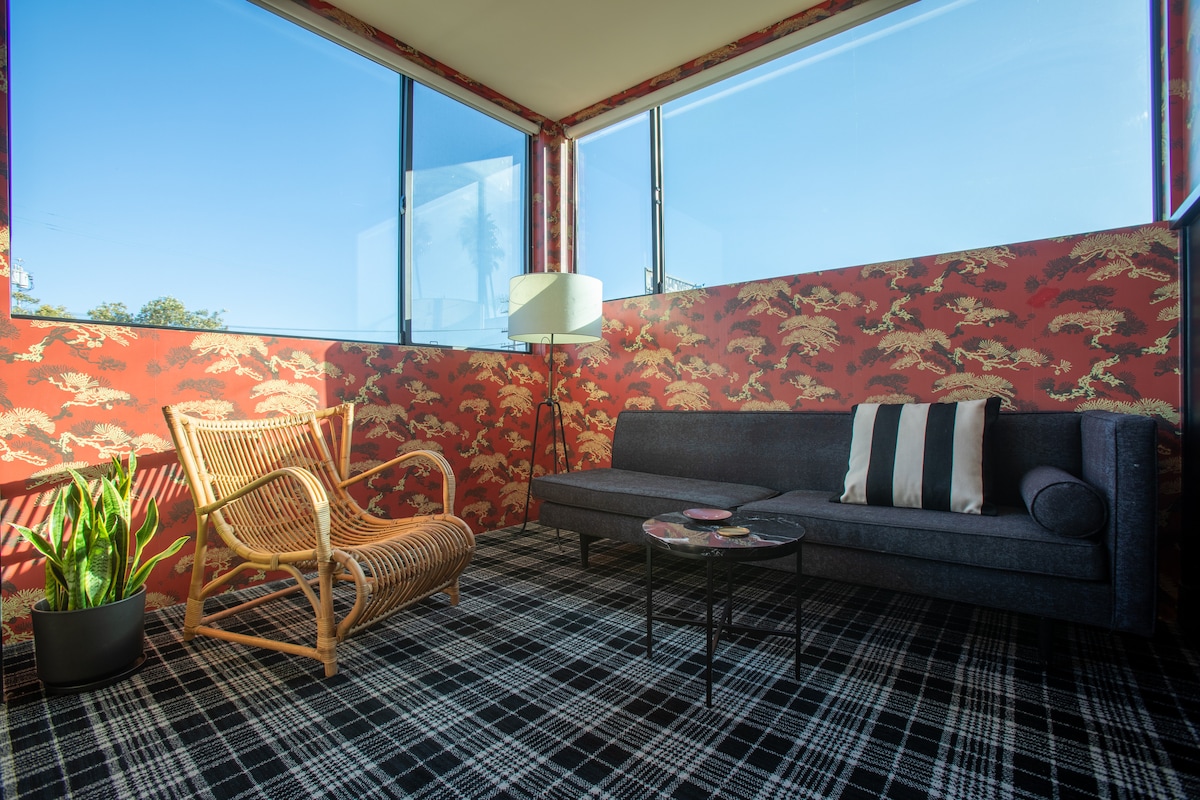 A cozy seating area is furnished with a dark sofa and a rattan chair, set against red patterned walls. A small coffee table sits in the center, and a potted plant adds a touch of greenery. Large windows allow plenty of natural light to fill the space.