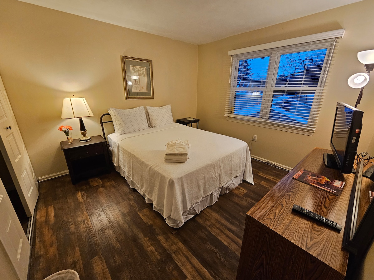 A bright sleeping area is furnished with a queen size bed, dressed in white linens. A bedside table with a lamp and a vase of flowers sits next to the bed. Natural light enters through two large windows with blinds, enhancing the room's neutral tones.