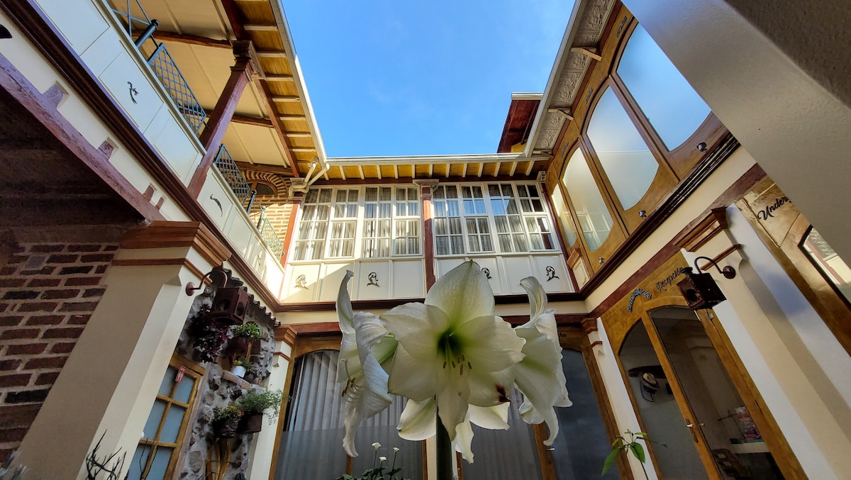 An open-air central patio is visible, framed by colonial-style corridors and large glass windows that invite natural light. A floral arrangement with white lilies adorns the foreground, while the sky above is clear and blue, enhancing the spacious atmosphere.