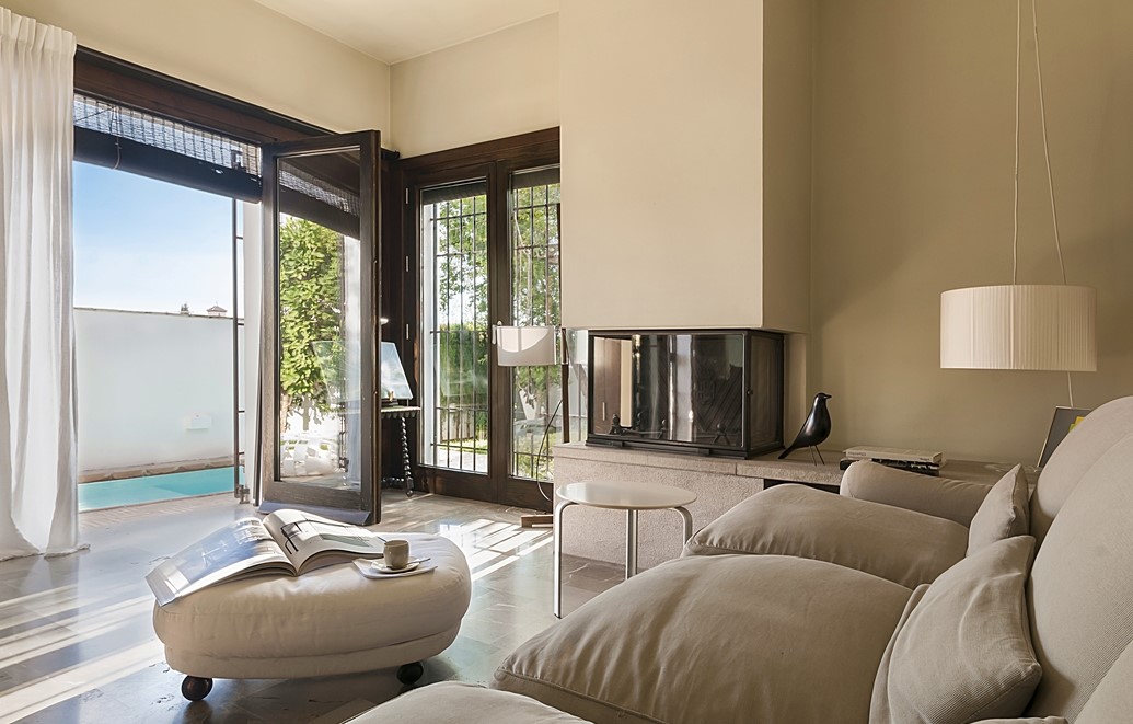 A bright living room showcases a comfortable seating area with two light-colored sofas facing a modern fireplace. Large glass doors open to a patio with views of the pool, allowing natural light to fill the space. A coffee table sits nearby with a book and cup.