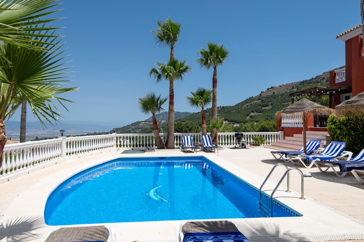 A private pool is surrounded by a spacious patio featuring lounge chairs. Palm trees provide shade alongside the pool, while the hillside landscape offers expansive views of the surrounding area. Clear blue skies enhance the relaxing atmosphere of the outdoor space.