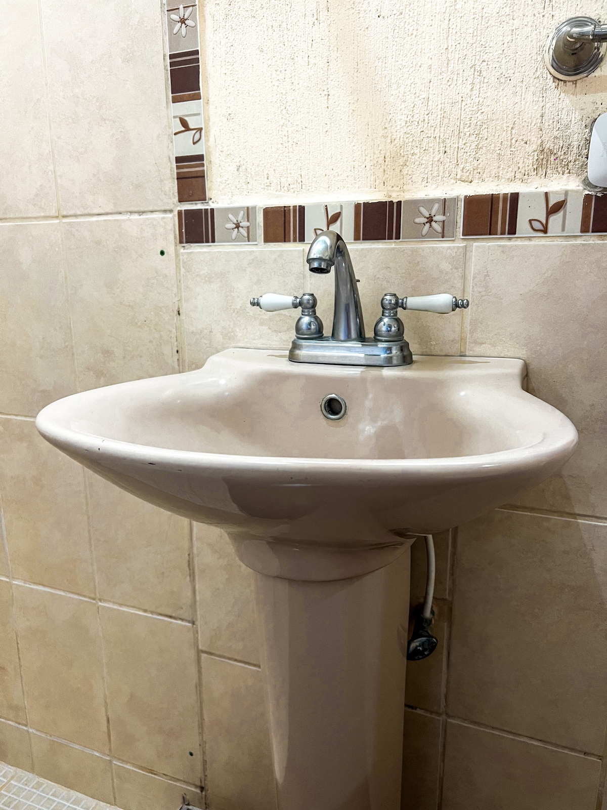 A clean sink is shown, featuring a smooth, rounded basin and a chrome faucet with white handles. The surrounding tiled wall displays a simple pattern with neutral tones, contributing to a functional and cozy bathroom space.