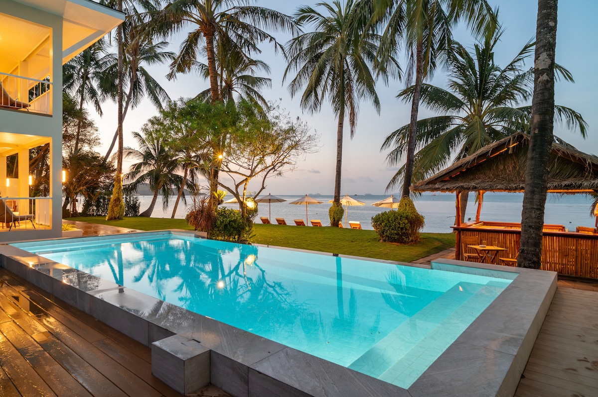 A serene infinity pool reflects the evening sky, surrounded by palm trees and plush loungers on a spacious deck. In the background, the beach and calm sea offer a tranquil backdrop, while a rustic bar area provides an inviting spot for refreshments.