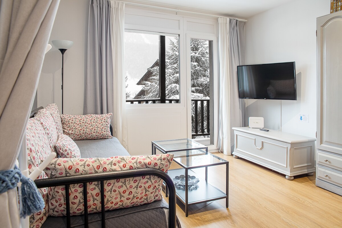 A cozy living area features a floral-patterned sofa bed and a glass coffee table. Natural light fills the space through large windows, showcasing views of trees and mountains. A mounted TV is positioned across from the seating area, with neutral wall colors providing a calm ambiance.