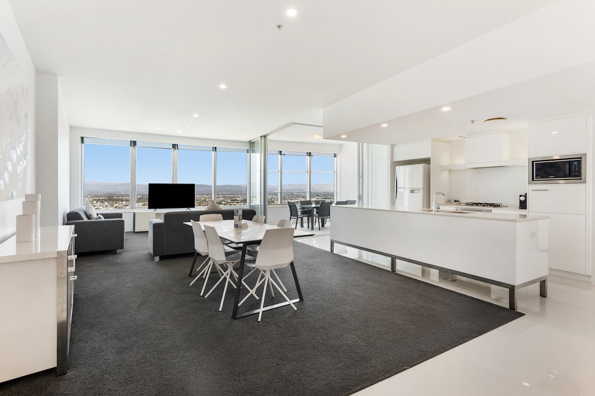 An open-plan living area is visible, featuring a modern kitchen with a gas stovetop and an adjoining dining space with a round table surrounded by chairs. Large windows provide sweeping views of the city and hinterland, filling the space with natural light.