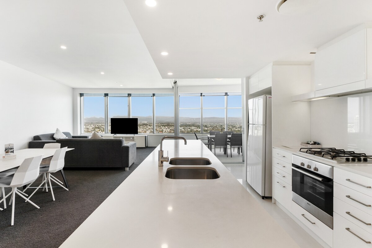 A bright and spacious living area is seen from the kitchen, featuring floor-to-ceiling windows that offer expansive views of the city and hinterland. Modern furnishings include a sleek kitchen with a gas stovetop and a comfortable sofa facing a flat-screen television.