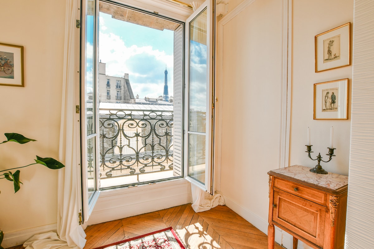 A bright corner of the room is showcased, featuring an open window with ornate ironwork. The Eiffel Tower can be seen in the background against a clear sky. A small wooden table stands beside the window, complemented by a potted plant on the floor.