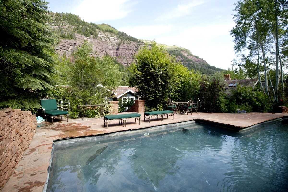 The outdoor swimming pool is surrounded by a natural stone patio, with relaxing lounge chairs positioned nearby. Lush greenery and mature trees frame the pool area, creating a serene environment. Towering mountain cliffs are visible in the background, providing a scenic backdrop.