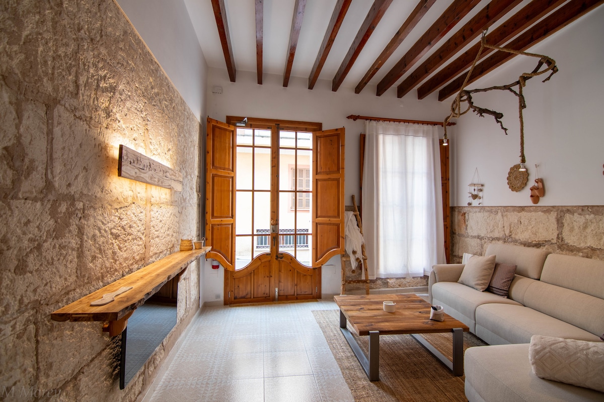 A spacious living area is illuminated by natural light streaming through large wooden windows. The walls showcase a blend of rustic stone and neutral tones. A comfortable sectional sofa faces a wooden coffee table, designed for relaxation and socializing.
