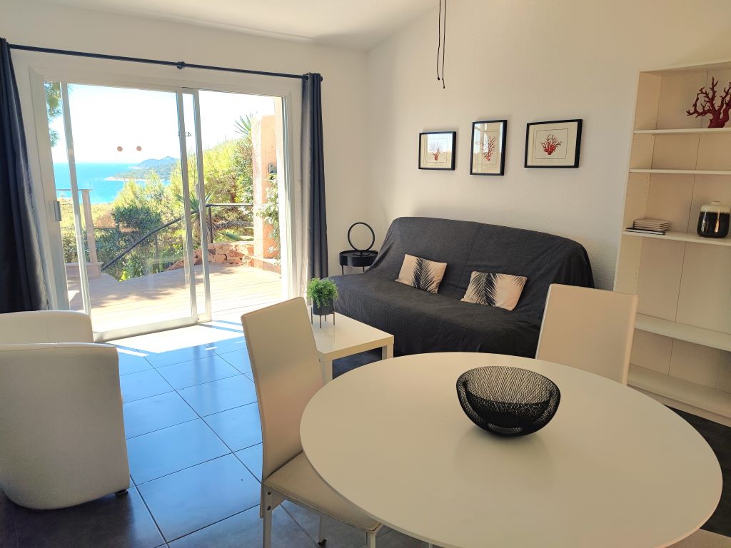 The living area presents a sofa bed, a round dining table, and modern chairs. Natural light enters through expansive glass doors, providing a view of the distant blue sea and lush greenery. Decorative wall art adds a touch of character to the space.