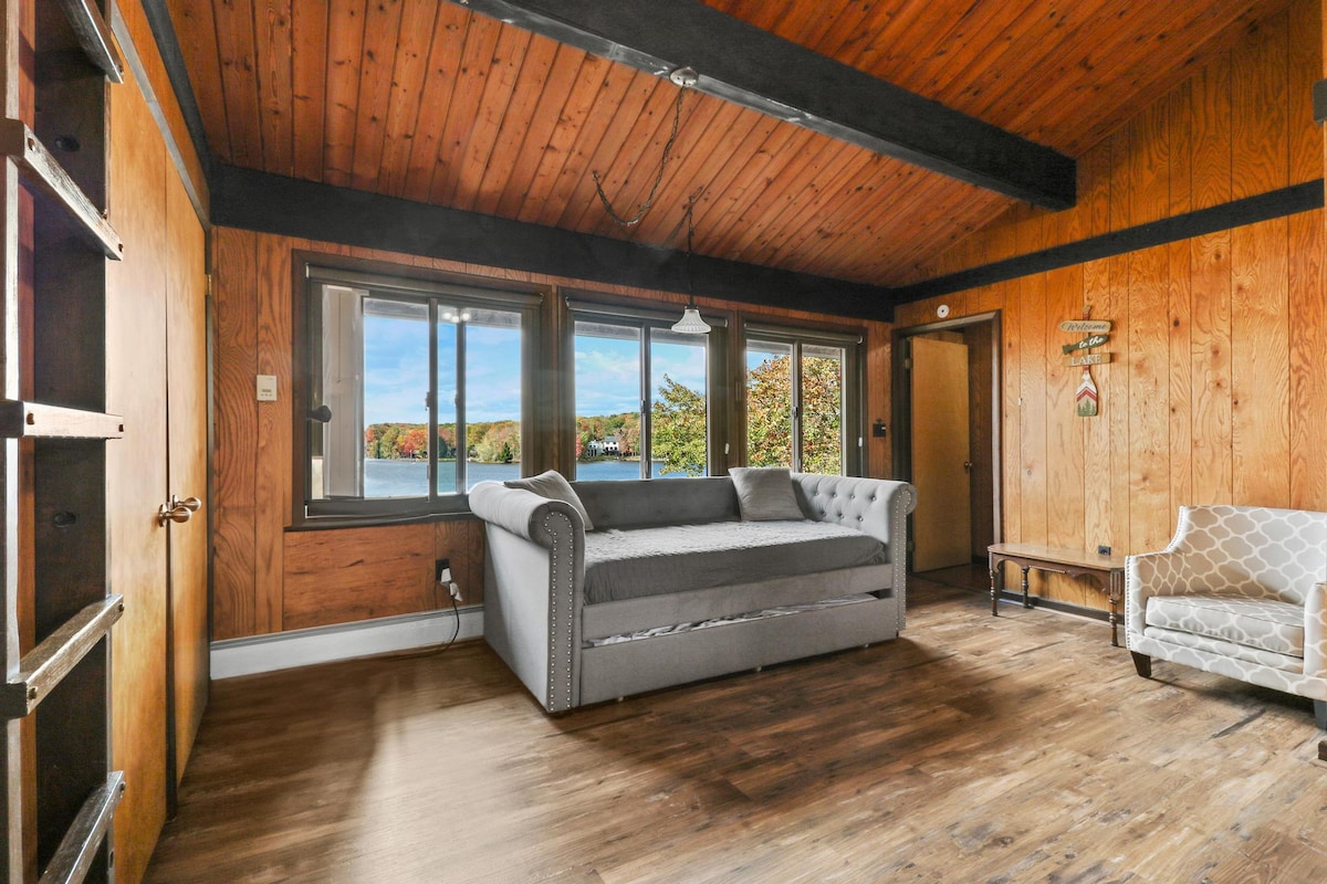 A cozy living area features wooden walls and a high ceiling with exposed beams. Large windows provide natural light and lake views. A plush sofa and an upholstered chair create inviting seating arrangements. The room's wood flooring complements the rustic atmosphere.
