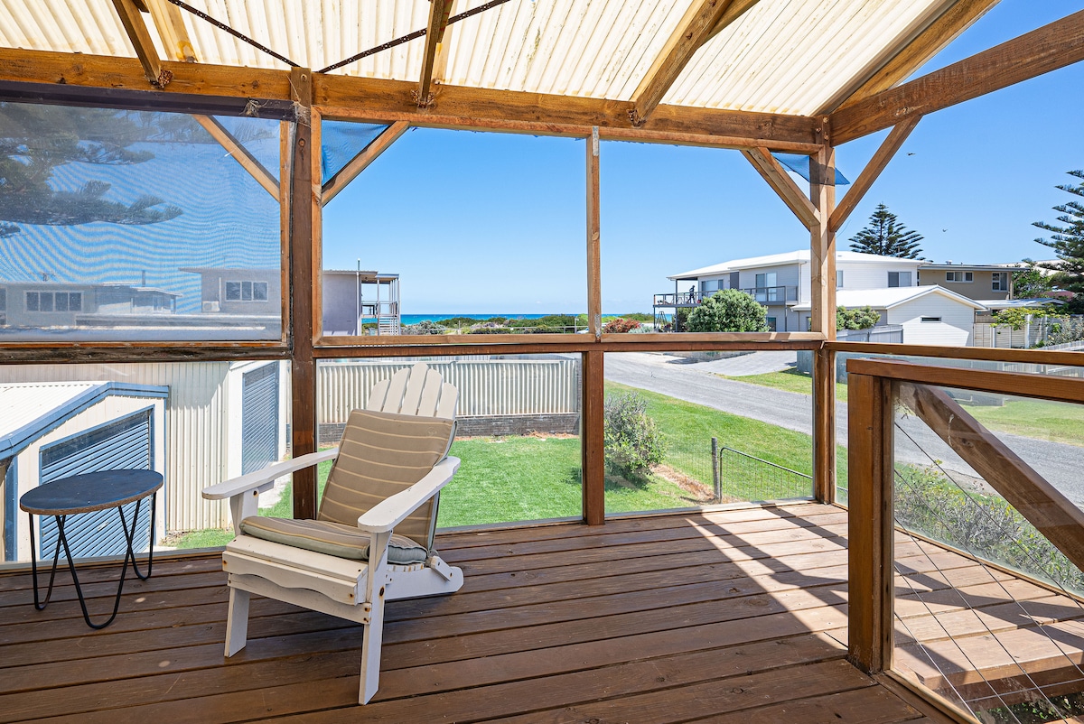 A covered deck area features a white adirondack chair and a small table, offering views of the nearby ocean and surrounding neighborhood. Natural wood beams support the structure, while large windows provide ample light and an inviting outdoor space.
