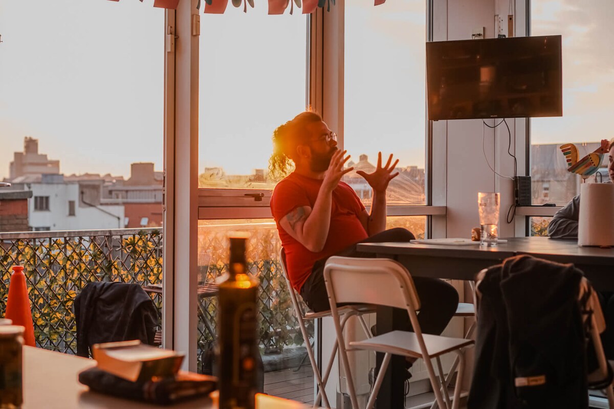 A dining area features a table surrounded by white folding chairs, with sunlight streaming through large glass doors. A television mounted on the wall displays a scenic view. The balcony outside shows vibrant greenery and overlooks city buildings.