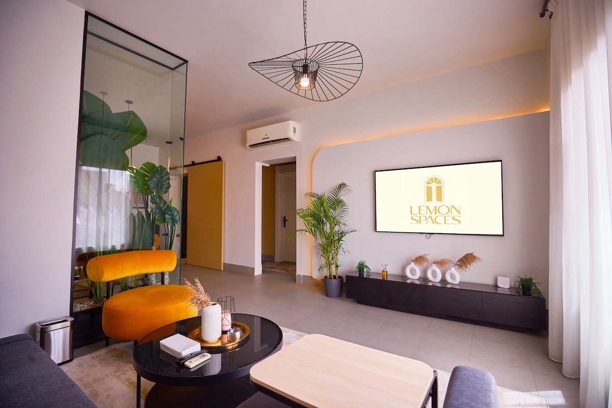 The living area features a modern design with a sleek black sideboard and a large smart TV mounted on the wall. A comfortable orange armchair complements a round glass coffee table, enhancing the space's cozy feel. Green plants add a touch of nature to the decor.